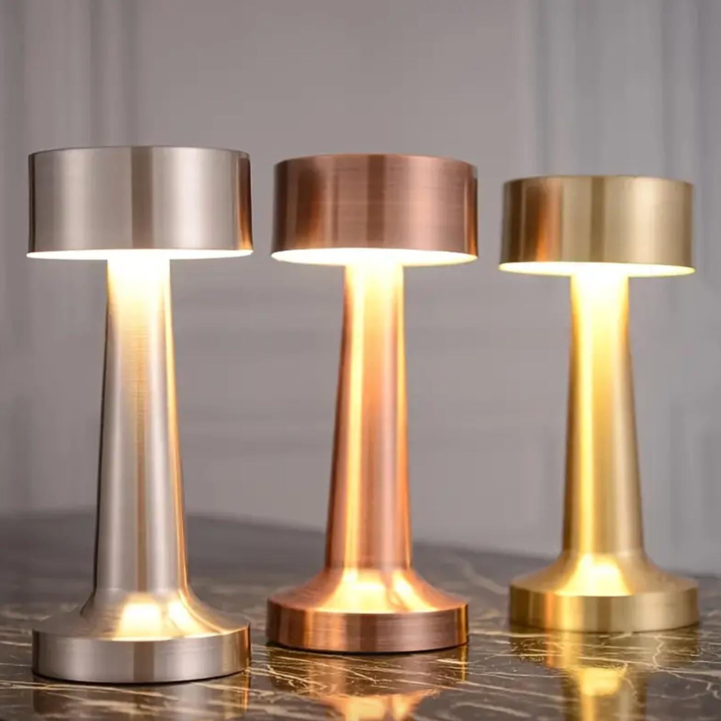 Three metallic table lamps in silver, bronze, and gold on a reflective surface with a neutral background.