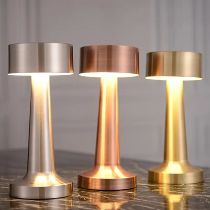 Three metallic table lamps in silver, bronze, and gold on a reflective surface with a neutral background.